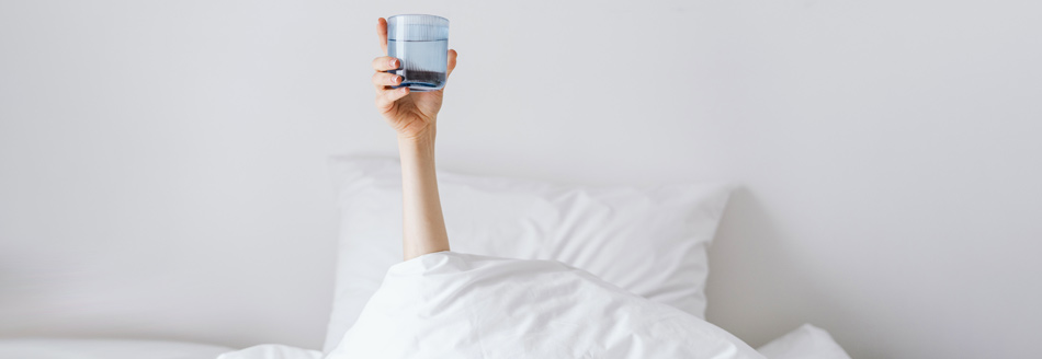 Was hilft gegen Kater? Eine Frau im Bett hält ein Wasserglas hoch