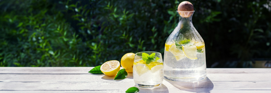 Eine Glaskaraffe ist mit kaltem Wasser, Eiswürfeln und Zitronenscheiben gefüllt und steht auf einem Tisch in der Sonne.