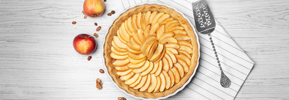 Ein fertig gebackener Apfelkuchen neben einigen Äpfeln und einem Tortenheber