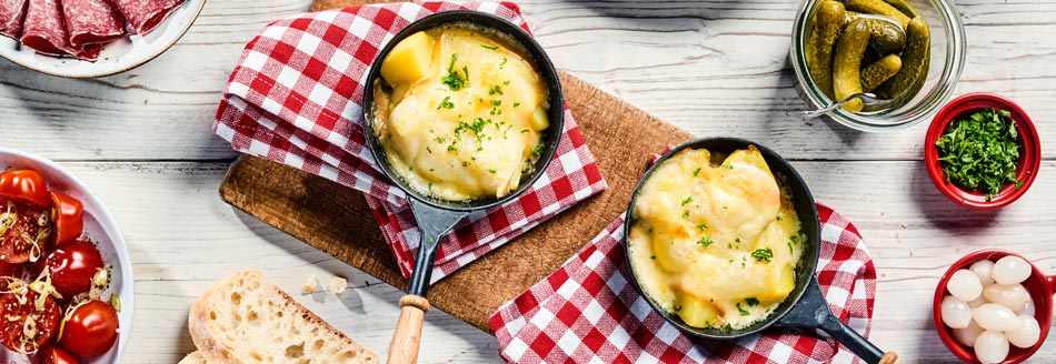 Welche ist die richtige Kartoffel für Raclette? Raclettepfannen liegen mit Zutaten auf einem Esstisch.